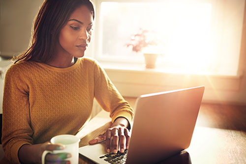 Woman at a desk, holding a hot drink, and using laptop to search for an insurance agent or adjuster by name or license number.